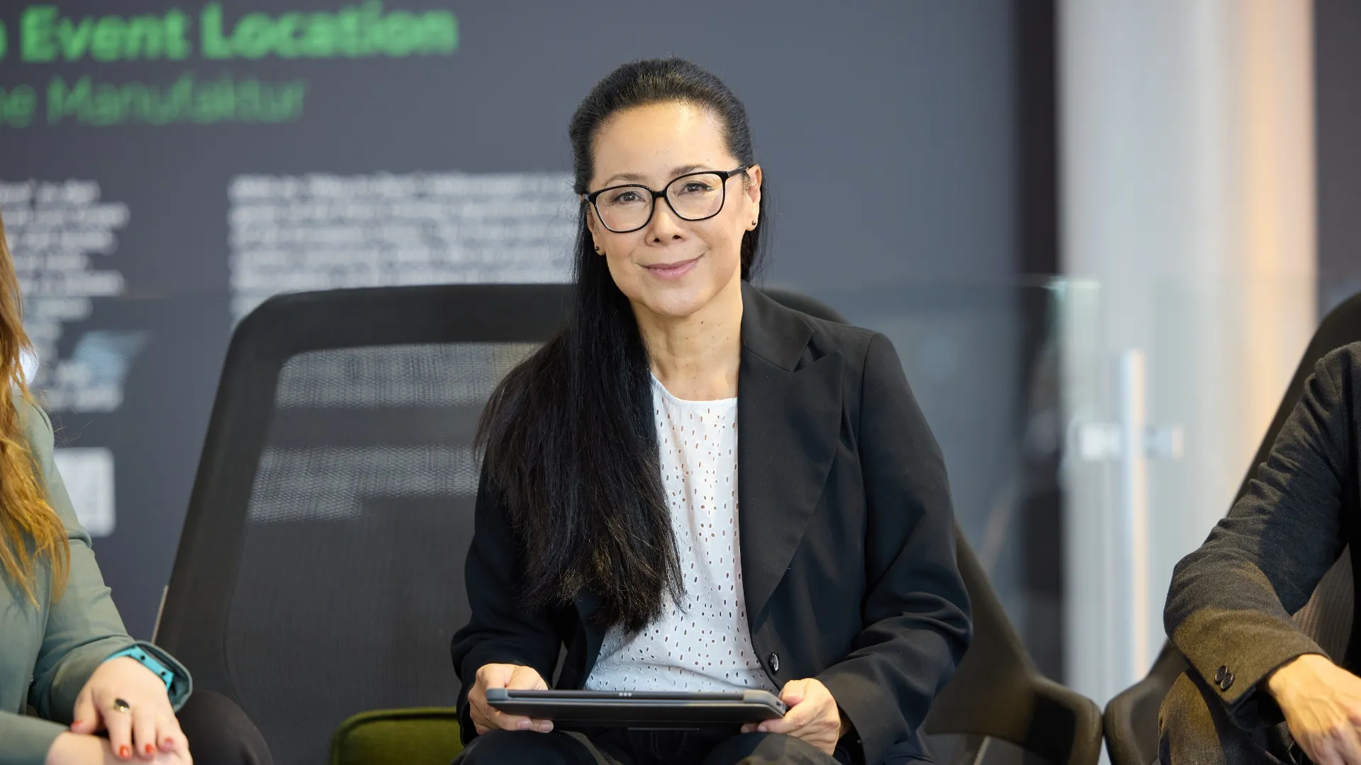 Ka Lam sitzt an einem Konferenztisch und hält ein Tablet Gerät in den Händen. Zwei Personen sitzen links und rechts zu ihr zugewandt. Im Hintergrund ist eine Wand.