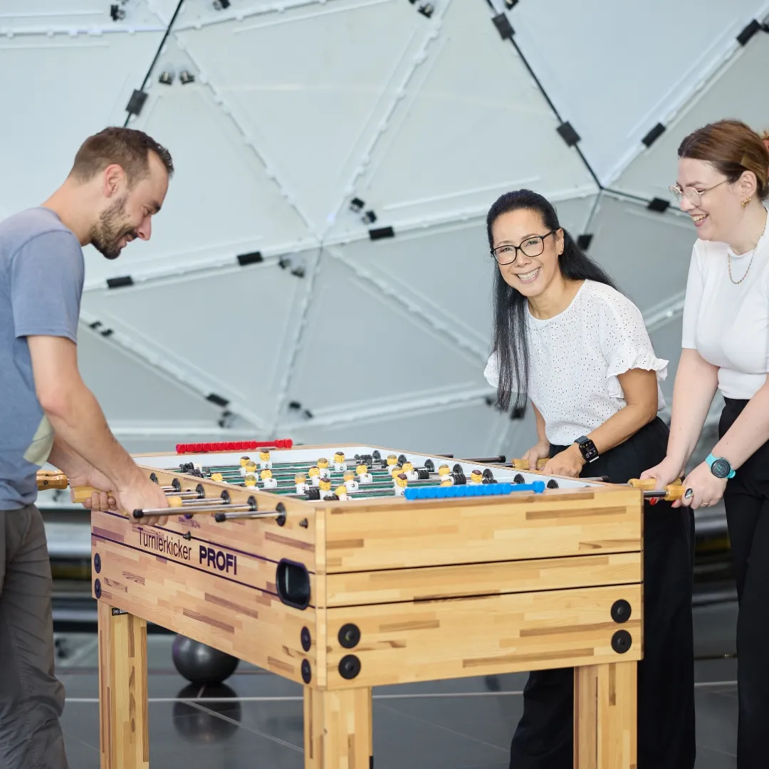 Drei Personen stehen um einem Tischfussball Spiel herum.